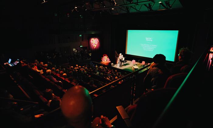 A view of an auditorium full with dimly audience members and a man standing on the stage, lit up and with a turquoise screen behind him. in the foregr